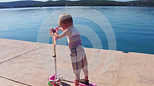 Little child learning to ride a scooter at seafront on sunny summer day. Cute preschooler boy riding a roller. Kids play