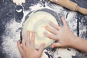 Little child making dough for backing. Kid`s hands, some flour, wheat dough and rolling-pin on the black table. Children hands ma