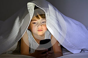 Little child play on smart phone in bed under the covers at nigh