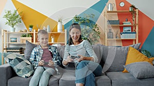 Little child playing video games with caring mother holding joysticks talking at home