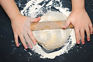 Little child preparing dough for backing. Kid`s hands, some flour, wheat dough and rolling-pin on the black table. Children hands