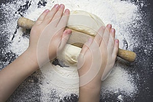 Little child preparing dough for backing. Kid`s hands, some flour, wheat dough and rolling-pin on the black table. Children hands