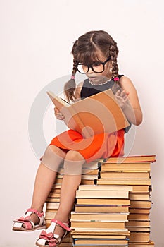 Little child reading book and in bewilderment covers mouth. Kid with glasses sitting on books.