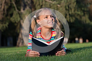 Little child reading book outdoors. Schoolgirl reading stories while relaxing green lawn. Cute pupil enjoy reading