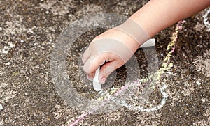 Little child`s hand, girl holding white chalk, drawing, writing on the concrete outside. School age kid drawing with chalk