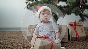 Little child in a Santa Claus hat holds a box with a gift. Sits on the floor near the Christmas tree