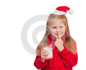 Little child in santa hat with glass of milk for Santa isolated. Beautiful little girl putting finger up to lips and ask silence