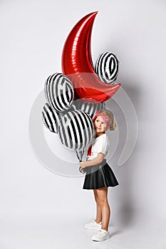 Little child in t-shirt, red headband, black skirt and sneakers. Holding balloons, posing sideways isolated on white. Full length