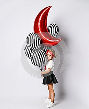 Little child in t-shirt, red headband, black skirt and sneakers. Holding balloons, posing sideways isolated on white. Full length