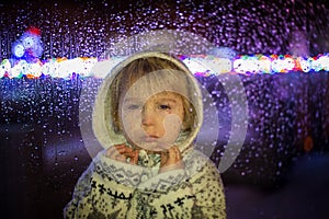 Little child, toddler boy, standing behind window with drops outdoors in the snow
