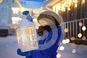 Little child is waiting Santa Claus on street of small town during snowfall on Christmas eve. Boy is holding large lantern with