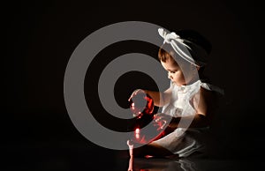 Little child in white headband and dress, barefoot. She playing with two red balls, sitting on floor. Twilight, black background.