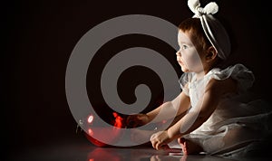 Little child in white headband and dress, barefoot. She playing with two red balls, sitting on floor. Twilight, black background