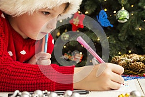 Little child is writing letter to Santa Claus at table. A kid in Santa hat is writing wishlist near the Christmas tree