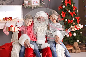 Little children with gift boxes sitting on Santa Claus` knees indoors