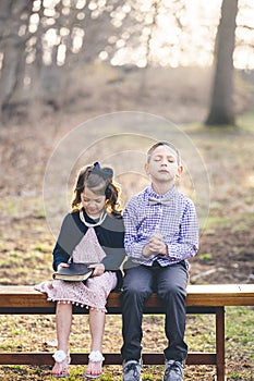 Little Christian boy praying and a girl holding the holy bible while sitting on a bench in a park