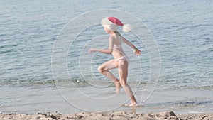 Little Christmas Girl in Santa Claus hat running at ocean sandy beach. celebrating Christmas traveling to warm countries