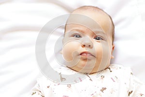 Little chubby newborn baby in a white bodysuit lying on the bed. Top view of a sad newborn baby on a white warm blanket.