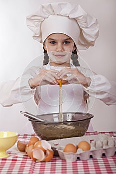 Little cook girl in a white apron breaks eggs in a deep dish