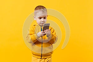 Little cool kid boy 3-4 years old in yellow clothes talking on mobile phone isolated on orange wall background, studio