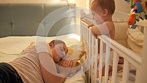 Little crying baby boy waking up his mother sleeping in bed at night. Concept of parenting, parent fatigue and children