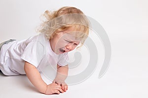 A little curly crying child in a clean white T-shirt is lying on the floor and crying.