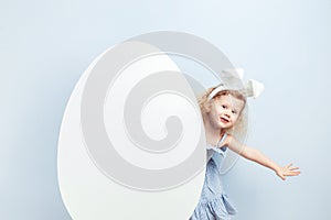 Little curly girl in the light-blue dress with bunny ears on her head looks out from behind a big white egg against a