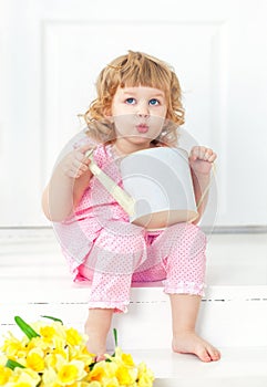 Little curly girl in pink dress and barefoot sits on a white porch and playing with children's watering can.