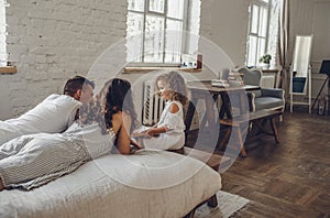 a little curly-haired girl is sitting next to the bed on which dad and mom are lying