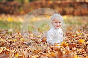 Little cute 3-4-year-old blonde girl sits on fallen leaves and smiles while walking in the autumn park