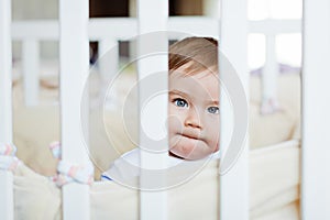 Little cute adorable little blond boy in a striped bodykit sitting with pursed lips behind bars white cot