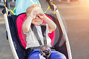 Little cute adorable toddler kid boy rubbing eyes with hands sitting in stroller on bright summer day outdoors. Tired