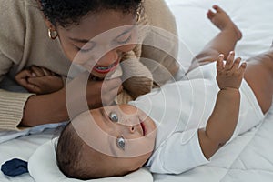 Little cute African American newborn baby lying on bed and looking smiling at camera while young mother is looking at infant