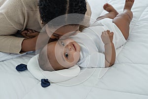 Little cute African American newborn baby lying on bed and looking smiling while young mother is kissing at infant cheek with love