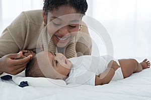Little cute African American newborn baby lying on bed and looking smiling at young mother who is touching infant`s head with love