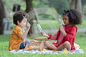 Little cute African children boy and girl sitting and have fun playing magnifying glass to read book while picnic at park together