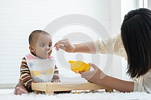 Little cute African newborn baby girl wear apron eating food and sitting on floor at home. Young mother feeding food by spoon