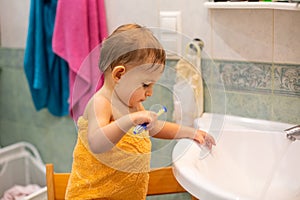 A little cute baby, 1,3 year old, is standing on a chair in the bathroom wrapped in an orange towel with a toothbrush