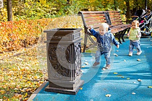 A little cute baby in blue clothes goes to a street bin in a park and throws garbage there and laughs while looking at