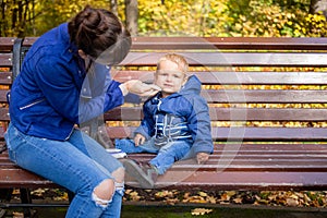 Little cute baby in blue clothes sits on a bench in the park, looks at the camera seriously, while Mom wipes his dirty
