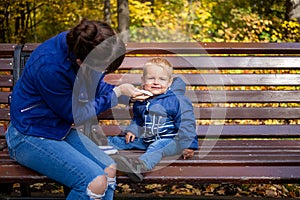 Little cute baby in blue clothes sits on a bench in the park, looks at the camera and smiles, while Mom wipes his dirty