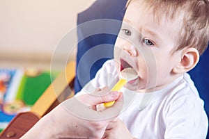Little cute baby eating on a chair in the kitchen. Mom feeds holding in hand a spoon of porridge