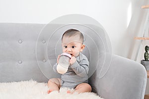 Little cute baby girl sitting in room on sofa drinking milk from bottle and smiling. Happy infant. Family people indoor Interior