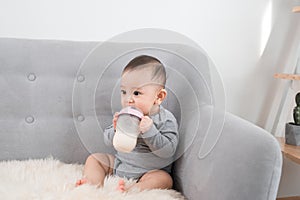 Little cute baby girl sitting in room on sofa drinking milk from bottle and smiling. Happy infant. Family people indoor Interior