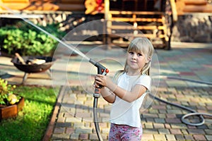 Little cute baby girl watering fresh green grass lawn mear house backyard on bright summer day. Child having fun playing with