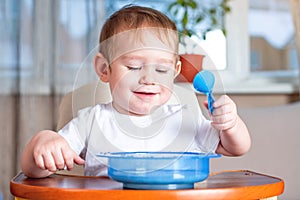 Little cute baby learning to eat with a spoon himself at the kids table in the kitchen. Healthy baby food