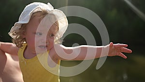 Little cute baby toddler girl blonde curls in yellow jumpsuit and white hat crying on mother's arms. Childish tantrum