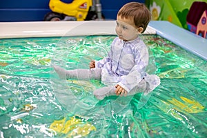 Little cute baby toddler girl in a dress sits in a toy pool on a children`s playground of day care play room, soft focus