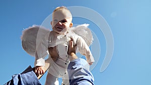 Little cute baby with white angel wings rised up in parent hands to blue sky