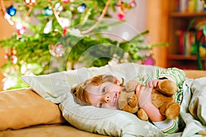 Little cute blond boy sleeping under Christmas tree and dreaming of Santa at home, indoors. Traditional Christian